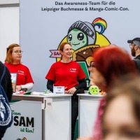 mehrere Personen vom Awareness Team SAM der Manga Comic Con 2025 stehen am Stand, Besucher:innen laufen daran vorbei mehrere Personen vom Awareness Team SAM der Manga Comic Con 2025 stehen am Stand, Besucher:innen laufen daran vorbei