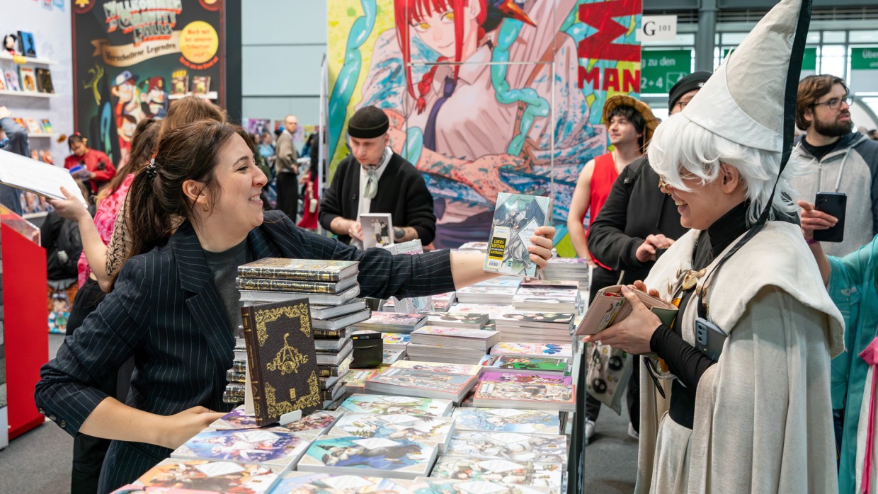 Ausstellerin überreicht Cosplayer:in ein Mangaheft über einen Verkaufstischer voller Manga auf der Manga Comic Con 2025 Ausstellerin überreicht Cosplayer:in ein Mangaheft über einen Verkaufstischer voller Manga auf der Manga Comic Con 2025